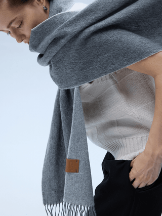 Gray fringed wool scarf draped over one shoulder of a white knit top, with a small leather label near the fringe