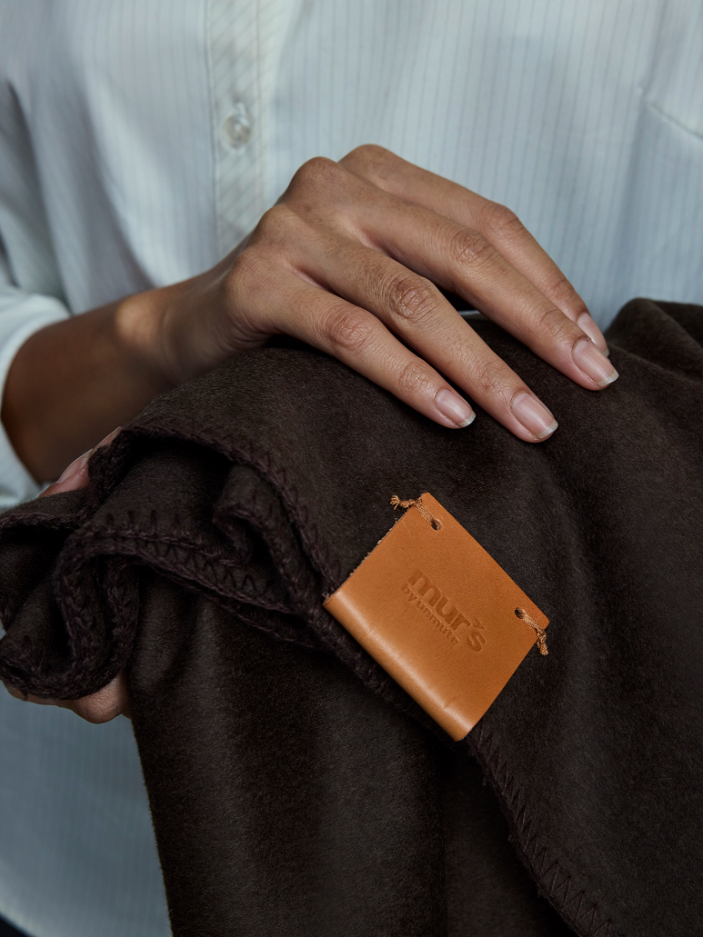 A close-up of a person holding a folded dark brown fleece blanket with a tan leather Murmurs tag attached. The person is wearing a light blue button-down shirt.