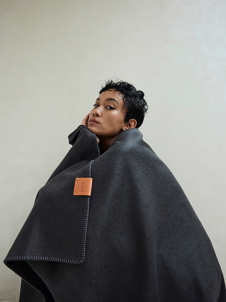 A person wrapped in a dark charcoal gray blanket or throw, photographed against a plain light beige wall. The throw has visible stitching along the edge and a small tan leather brand patch.