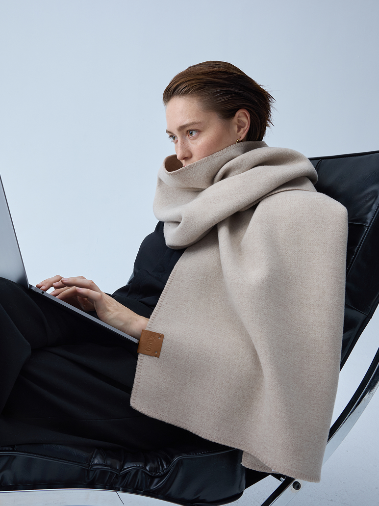 A woman sits in a black chair using a laptop, wrapped in a thick beige scarf with a small brown leather brand tab.