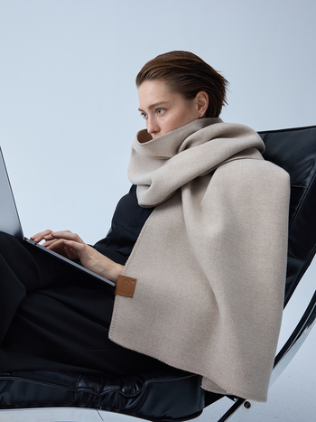 A woman sits in a black chair using a laptop, wrapped in a thick beige scarf with a small brown leather brand tab.