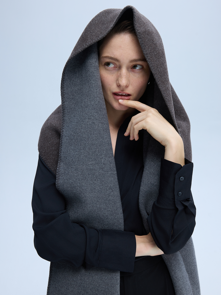 A model wearing a dark gray woolen hooded cape over a black long-sleeve top, posed against a solid light blue-gray studio background.