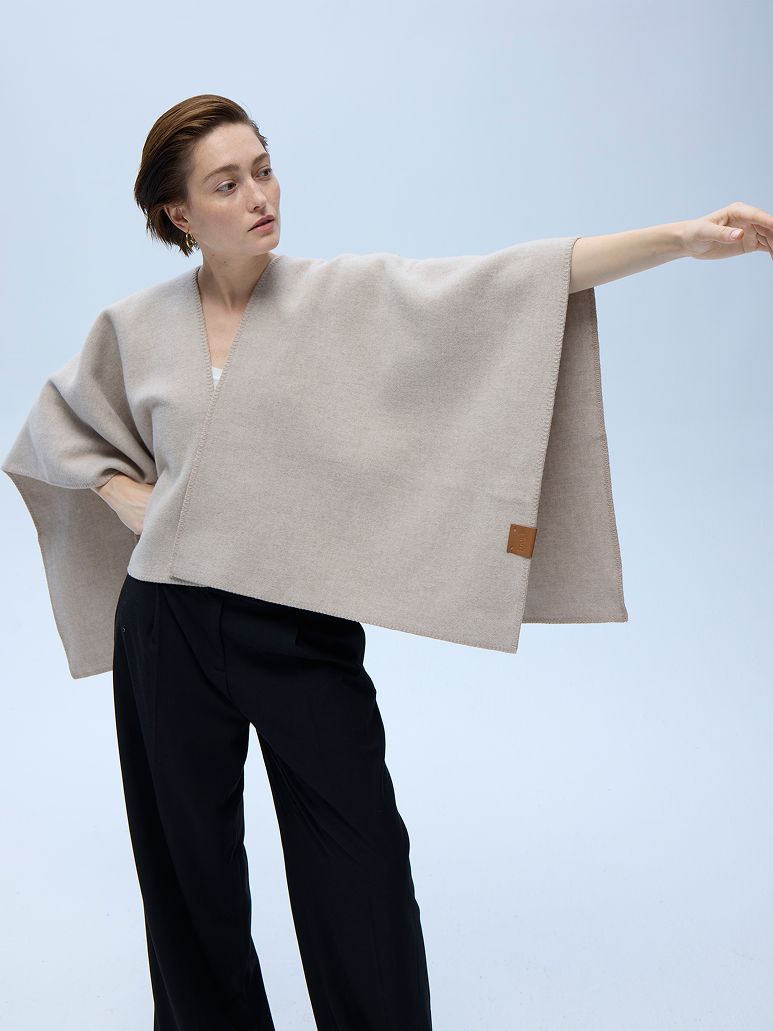A woman modeling a light beige open-front poncho-style wrap with a small brown leather patch, paired with black trousers, against a light blue studio background.
