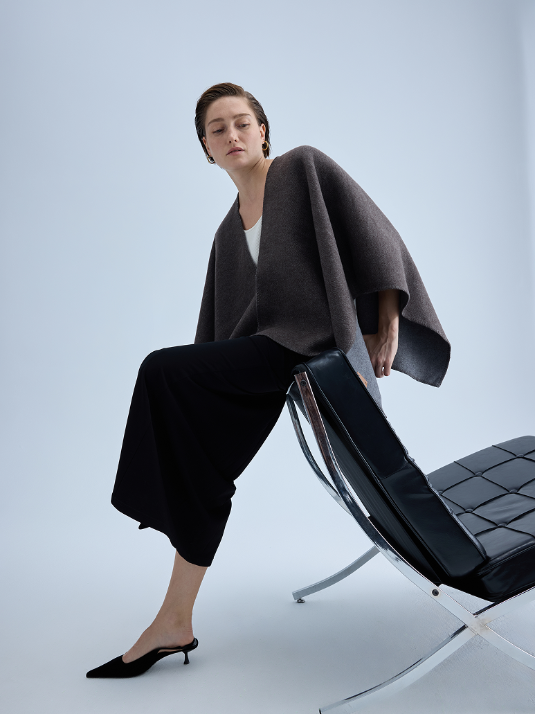 A model wearing a dark brown draped poncho-style cardigan, black wide-leg cropped pants, and black pointed mule heels, sitting on a black leather modern chair against a light gray studio background.