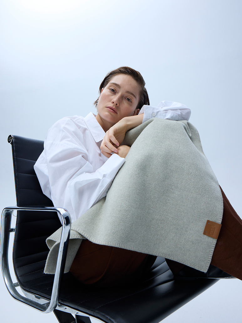 A woman reclining in a black office chair, wrapped in a light sage green knit throw blanket with a small brown leather brand tab. She is wearing an oversized white button-down shirt and brown trousers against a clean, light gray studio background.