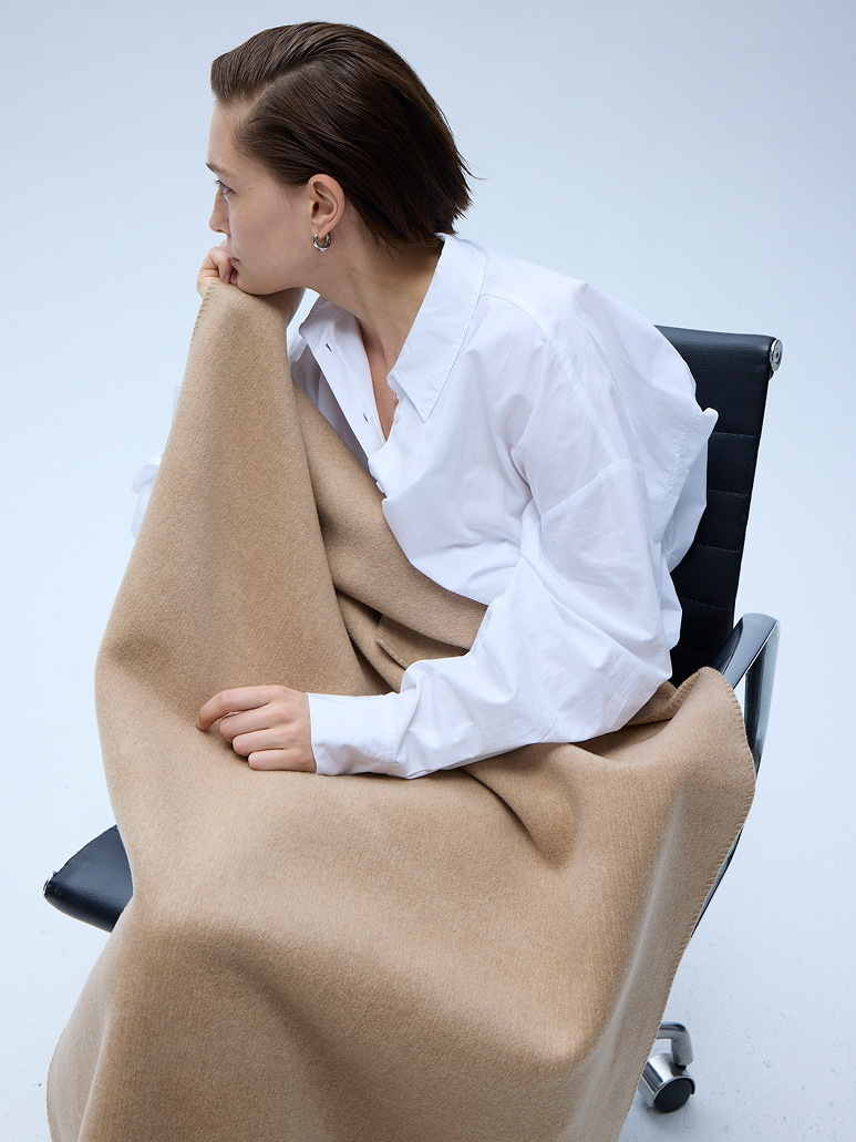 A woman with short brown hair sits in a black office chair, wrapped in a beige knit blanket. She is wearing an oversized white button-down shirt.