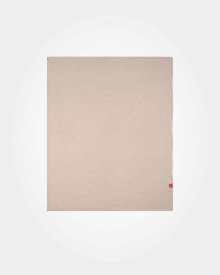 A minimalist, light beige rectangular fabric item, likely a blanket, throw, or scarf, featuring a tiny brown leather tag on the bottom right corner, displayed against a clean white background.
