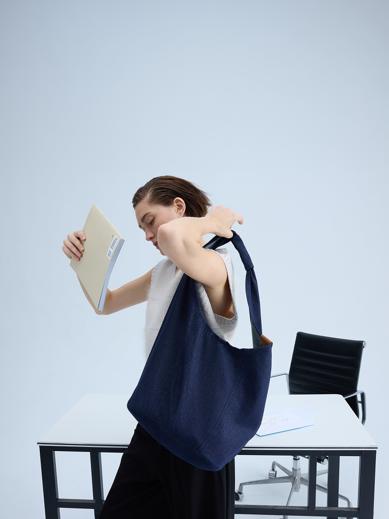 A woman in a white sleeveless top and black skirt carries a large navy blue textured fabric tote bag over her shoulder in an office setting with a desk and chair.