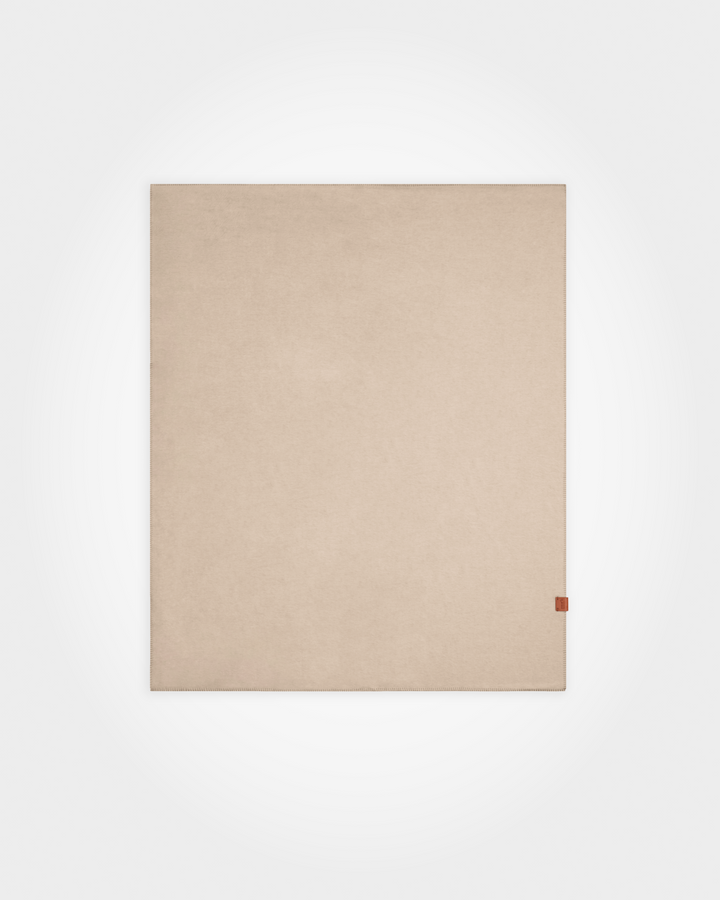 A minimalist beige rectangular mat or rug with a small brown leather tag on the bottom right corner, displayed against a clean white background.