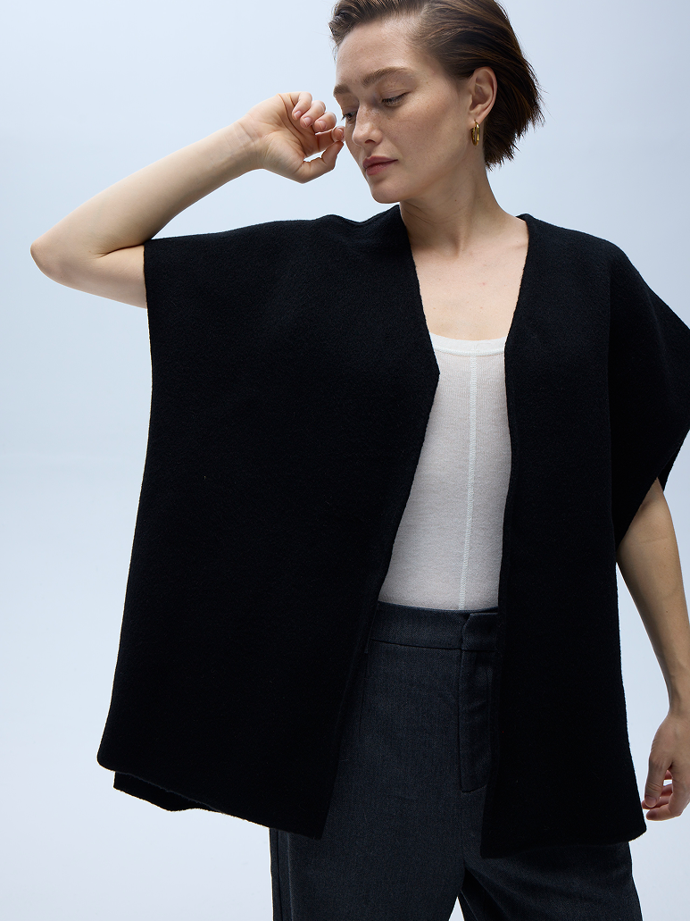 A woman modeling a black open-front cape-style cardigan or poncho over a white top and dark trousers, photographed against a clean light gray studio background.
