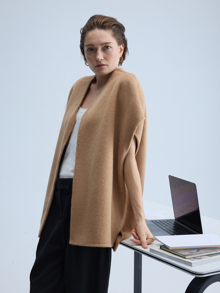 A woman models a camel-colored open-front cardigan or poncho over a white top and black pants, standing next to a desk with a laptop in a clean studio setting.