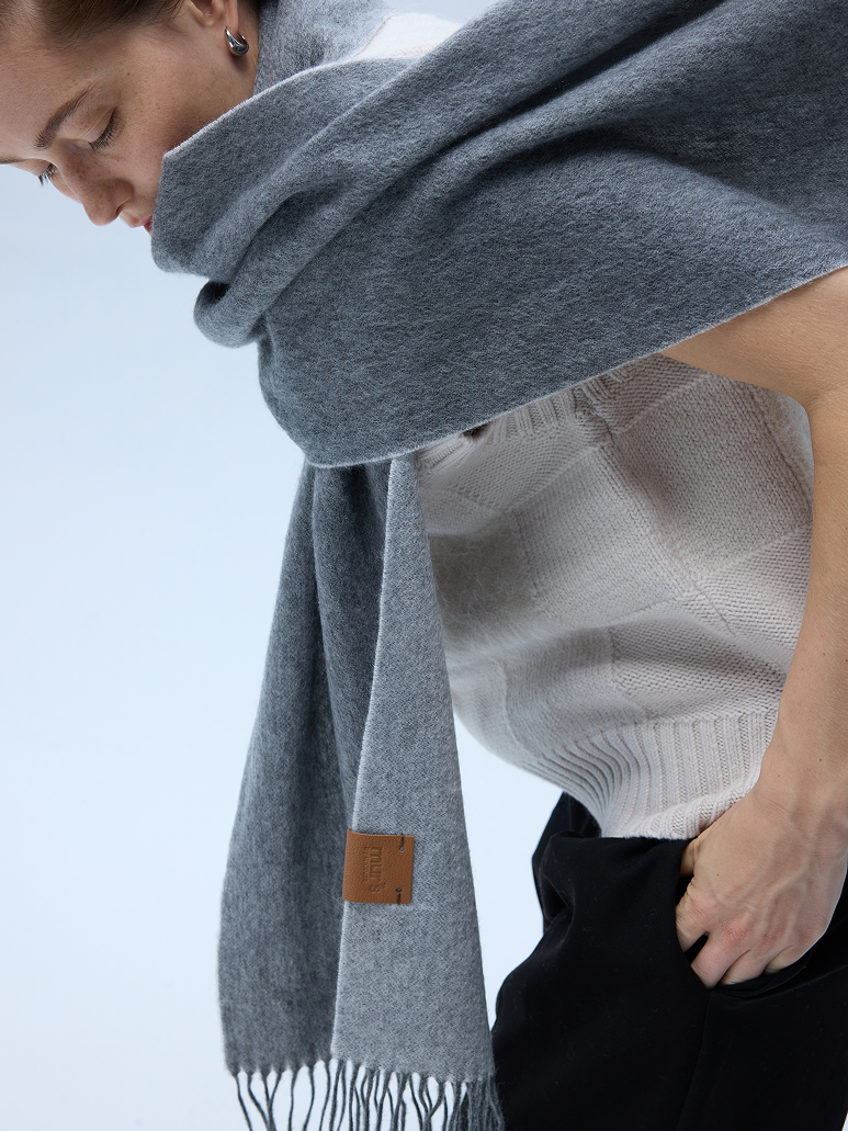 A close-up lifestyle shot of a woman wearing a soft, heather gray wool scarf wrapped around her neck and shoulders, paired with a cream knit sweater and black trousers. A small brown leather brand tag is visible on the scarf.