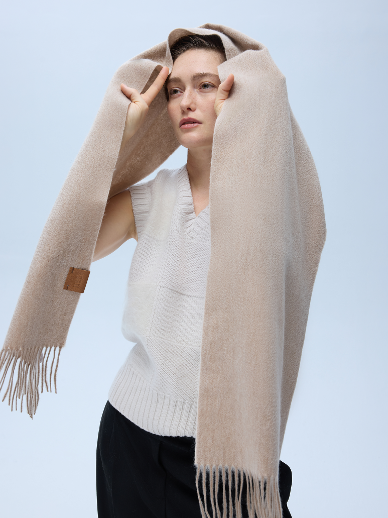 A woman modeling a soft, light beige oversized scarf with fringe, draped over her head and shoulders, paired with a white knit sweater vest and black pants against a light blue studio background.
