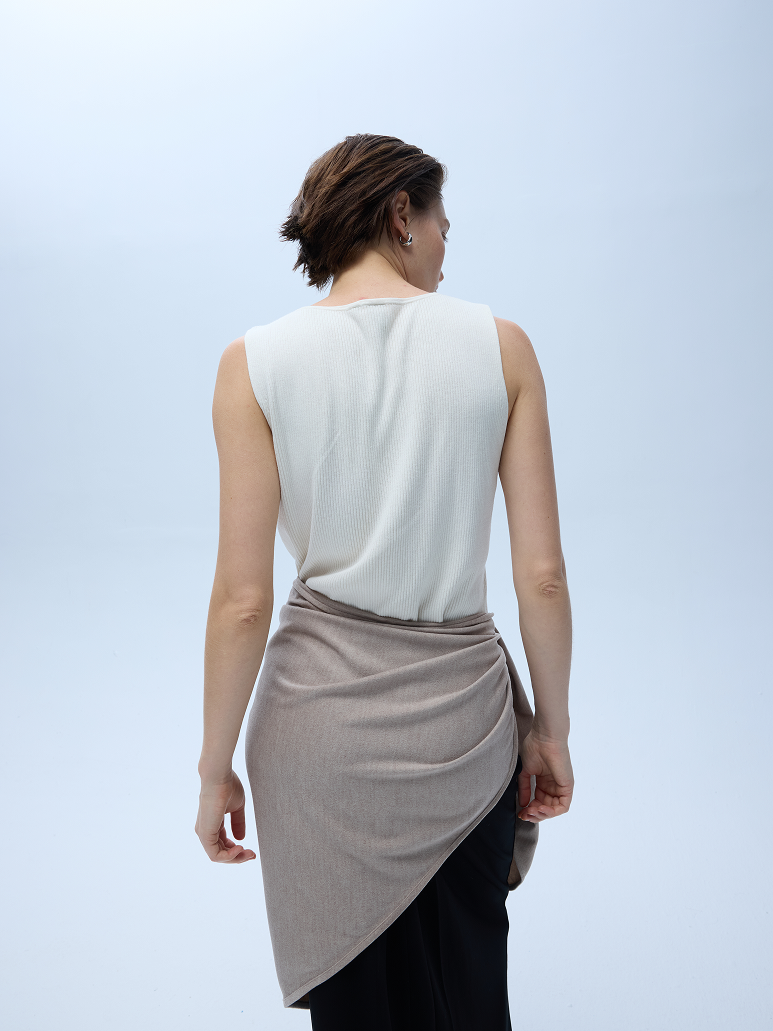 A woman shown from behind wearing a sleeveless white knit top and a draped taupe wrap skirt over black pants, against a light blue studio background.