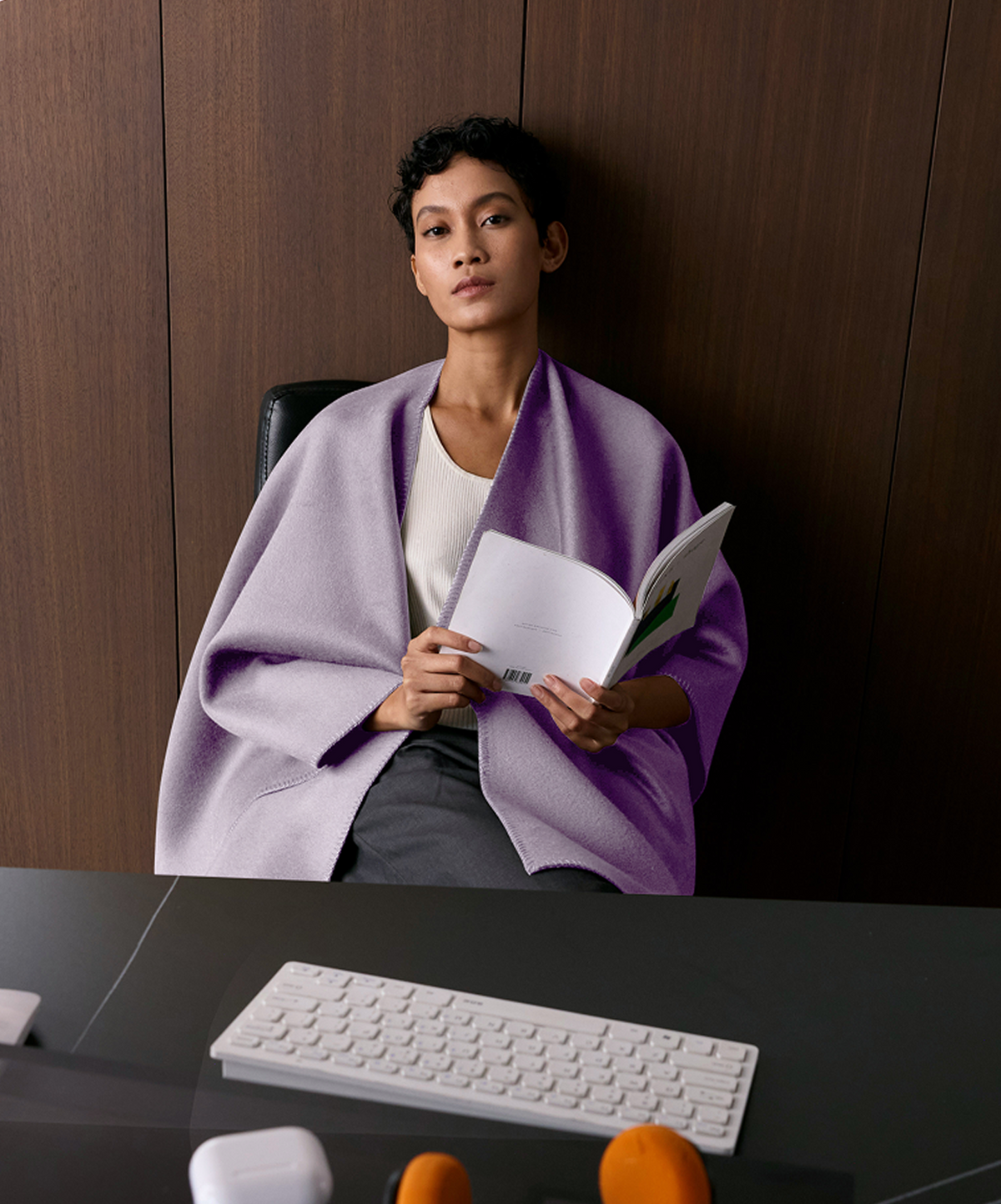 A woman sits in a dark office chair wrapped in a light purple blanket, holding an open magazine. She is wearing a white top and dark gray pants.