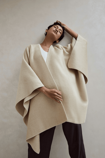 Woman wearing the Murmur wearable blanket wrap in Sand Dune cream, over a white knit and dark trousers | Murmurs by Unmute