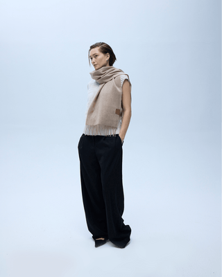 Woman wearing the Murmurs Whisper long cashmere scarf in Wheat Haze beige draped at the neck, fringed ends and leather label visible, over black wide-leg trousers | Murmurs by Unmute